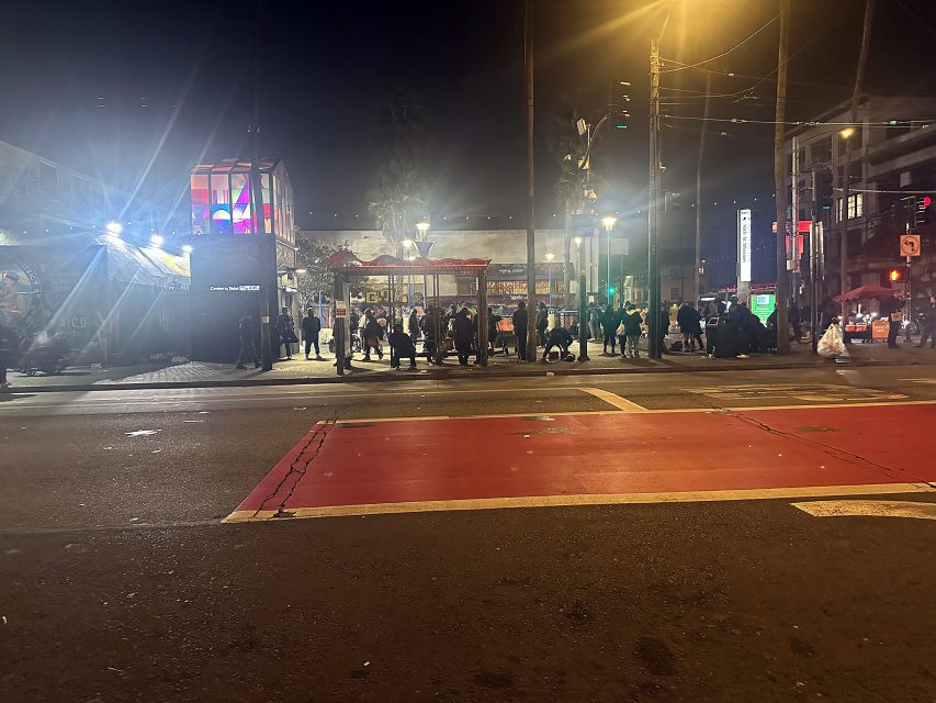 A group of people gather on a city sidewalk at night, across a wide street with red transit lanes, under streetlights and illuminated signs.