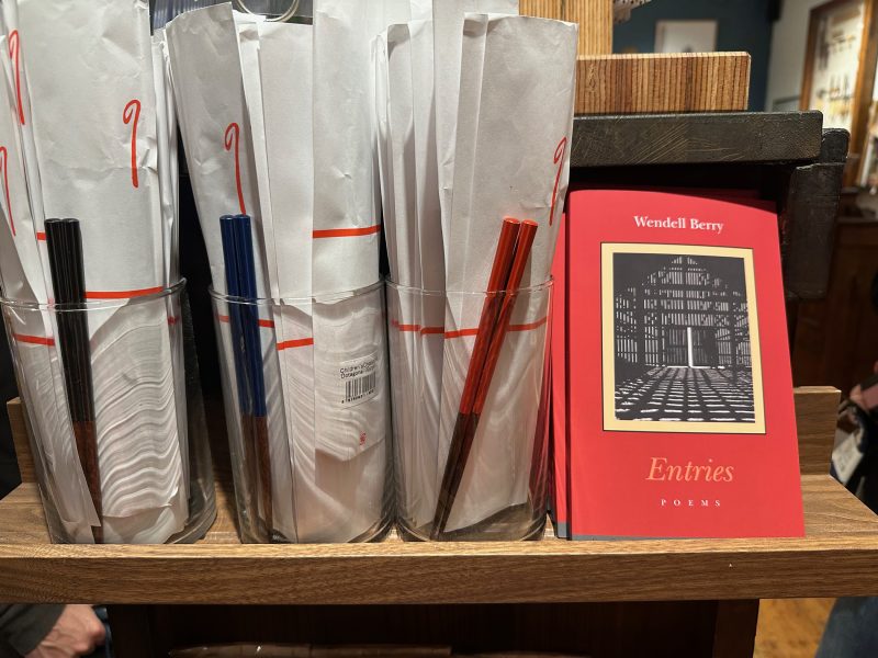 Rolled papers with pens in glass containers sit on a shelf next to a red book titled "Entries: Poems" by Wendell Berry.