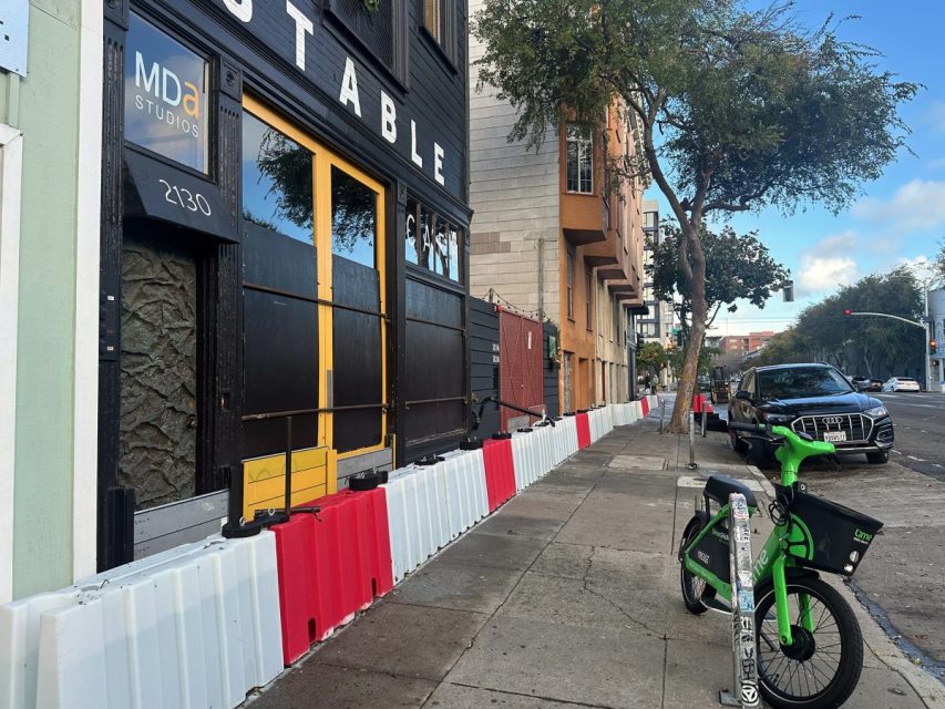 A sidewalk with red and white barriers, Lime e-bike and e-scooter parked, and a row of buildings and cars lining the street.