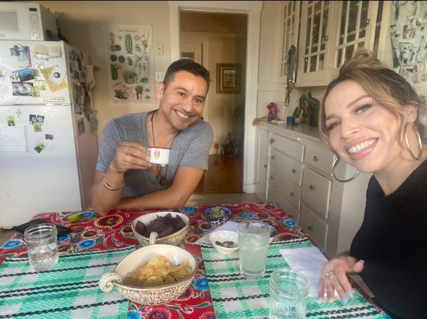 Two people sit at a colorful table with food and drinks in a kitchen, smiling at the camera.