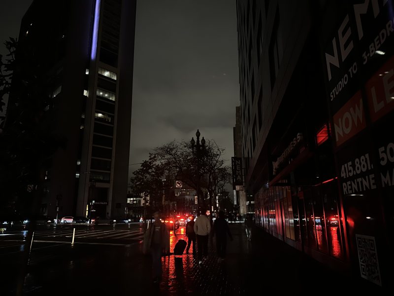A dark city street at night with wet pavement, illuminated by red lights from cars and building signs, and a few people walking on the sidewalk.