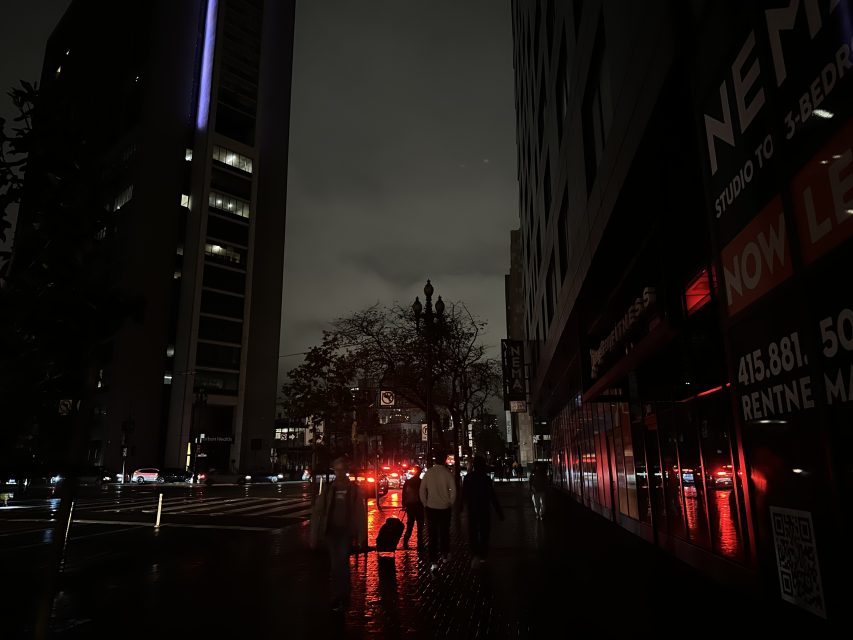 A dimly lit city street at night with people walking on the sidewalk, illuminated by red lights reflecting off wet pavement and buildings.
