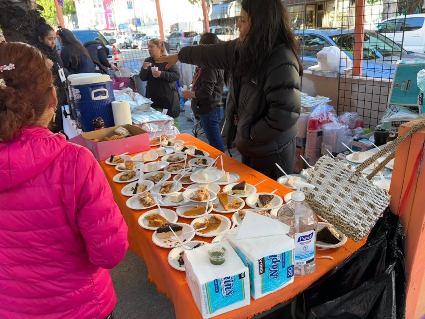 People stand by an outdoor table covered with plates of food samples, napkins, hand sanitizer, and other items at a street market.