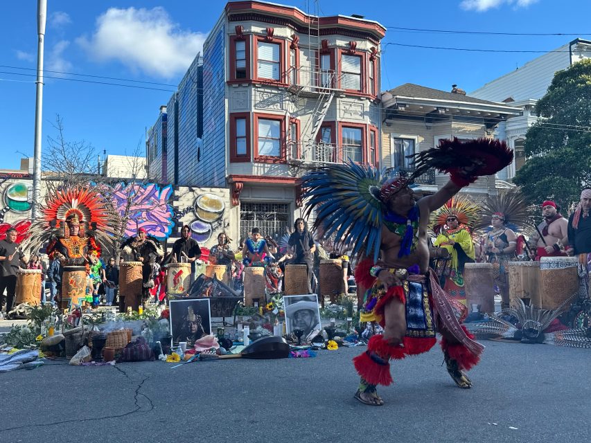 A group of people in traditional Aztec attire perform a ceremonial dance on a city street, with a colorful mural and offerings in the background.