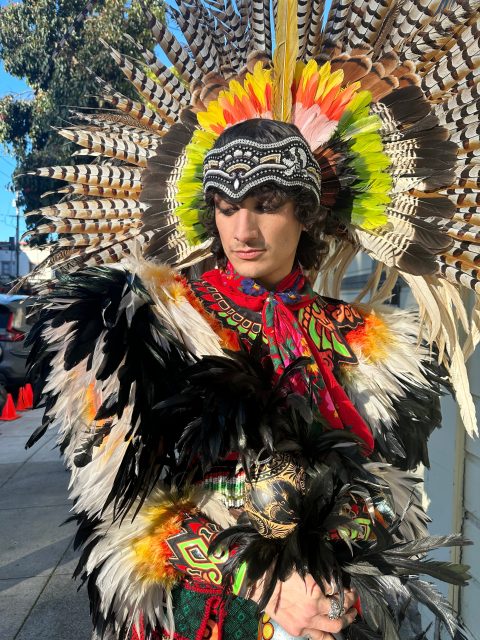 A person wearing a colorful traditional feathered headdress and embroidered clothing stands outside on a sunny day, looking down with hands clasped.