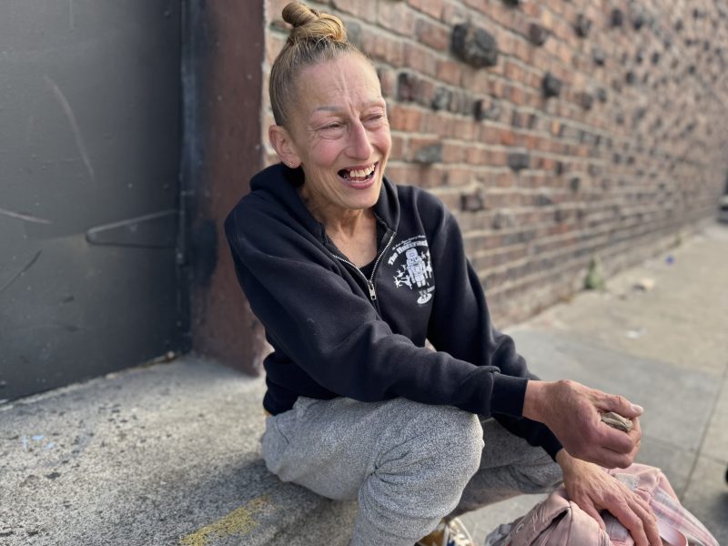 A woman with a topknot hairstyle sits on a sidewalk by a brick wall, smiling and wearing a black hoodie and gray sweatpants.