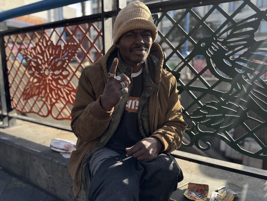 A man sits outdoors on a concrete ledge, wearing a brown jacket and beige knit cap, holding up one finger. Decorative metal fencing with red and black designs is behind him.