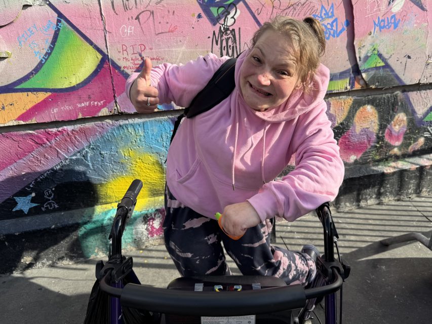 Person in a pink hoodie smiles and gives a thumbs up while using a walker, standing in front of a colorful graffiti-covered wall.