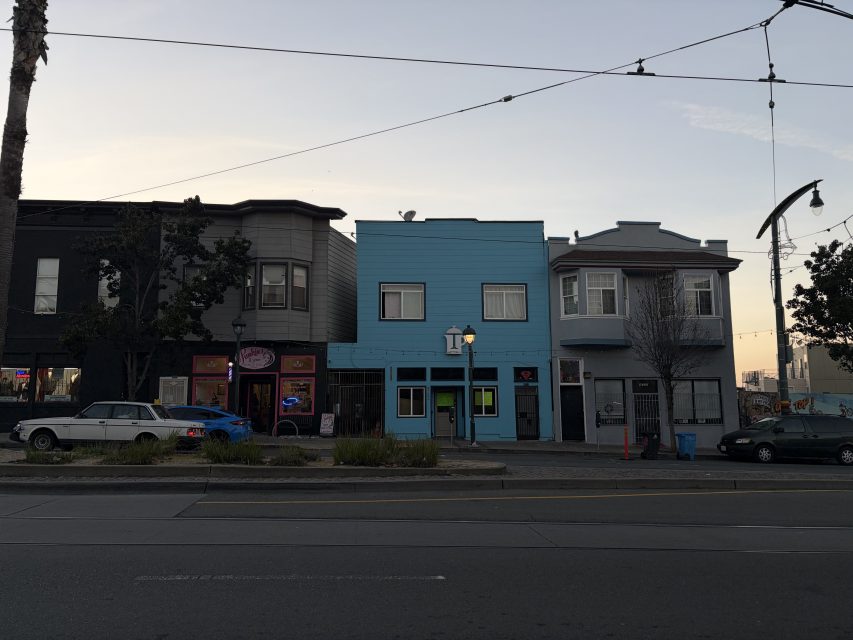 Three storefronts, including a bright blue building, line a quiet street at dusk with a few parked cars and overhead wires visible.