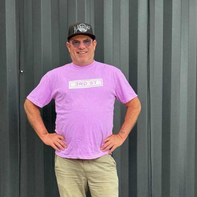 A man wearing a purple "3rd ST" t-shirt, khaki pants, and a black cap stands smiling in front of a gray corrugated metal wall, embodying the Bayview Buzz.