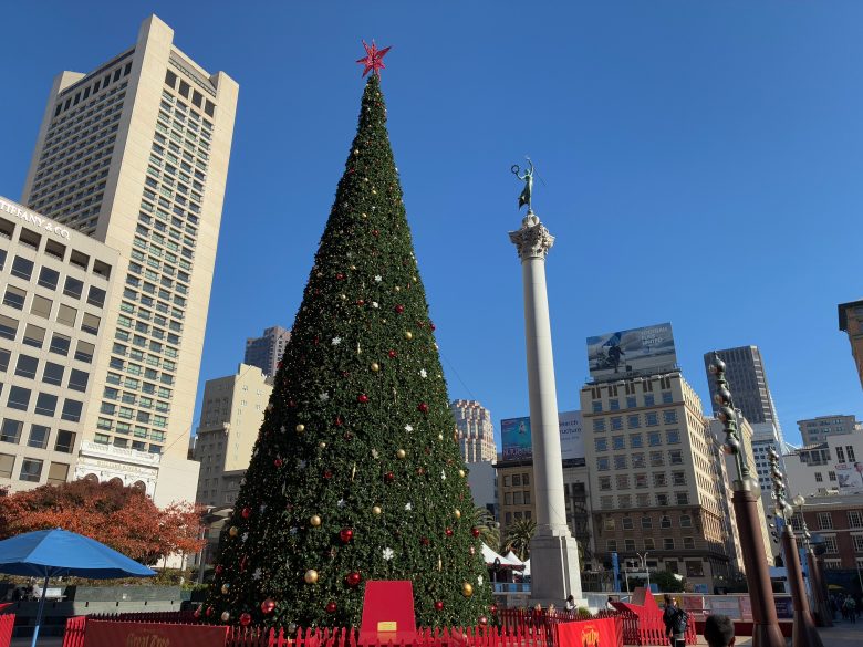 A large decorated Christmas tree stands in an urban square surrounded by tall buildings and a column with a statue on top under a clear blue sky.