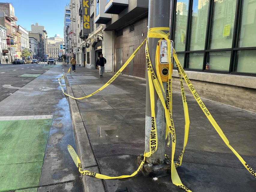 Yellow caution tape is wrapped around a pole with a pedestrian crossing button on a city sidewalk; a few people walk in the distance.