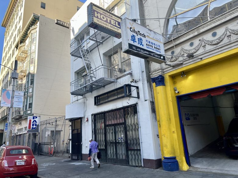 A person walks past a white building with a "HOTEL" sign, a garage entrance, and multiple business signs on an urban street.
