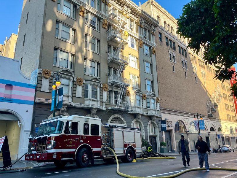 A San Francisco fire truck is parked outside a multi-story building with a hose extended, while two people walk by on the sidewalk.