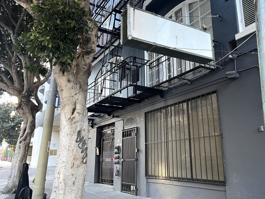 A dark gray building with barred windows, a metal door, a blank white hanging sign, and fire escape stairs above the entrance. Trees line the sidewalk in front.