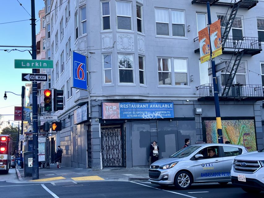 A corner building at Larkin Street with a Motel 6 sign, a "Restaurant Available" sign, and a taxi turning at the intersection. Pedestrians are on the sidewalk.