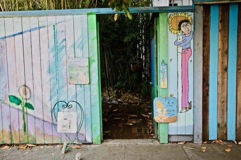 A wooden fence with colorful paintings, including a tall figure with a sunflower hat, surrounds an open gate leading to a path lined with plants. Signs and drawings are displayed on the fence.