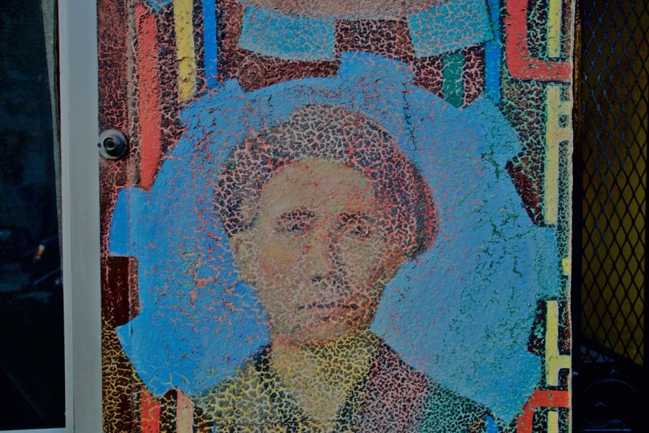 A textured mural depicts a person’s face within a blue circle, surrounded by vibrant, geometric patterns on a weathered surface.
