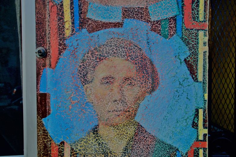 A weathered mural depicts a person's face with short hair, surrounded by blue and red geometric shapes, next to a door and a mesh panel.