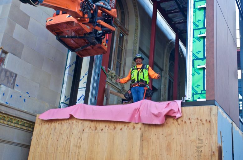 Construction worker in safety gear stands on a wooden platform near a building facade, with machinery and insulation visible in the background. Photo by Walter Mackins