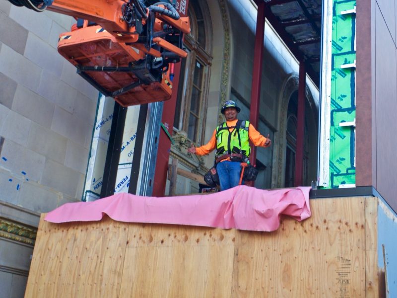 Construction worker in safety gear stands on a platform next to a building under renovation, with machinery and unfinished walls visible.