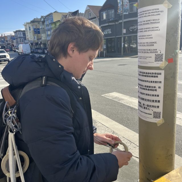 A person with a backpack tapes a printed notice onto a yellow pole at a street corner on a sunny day.
