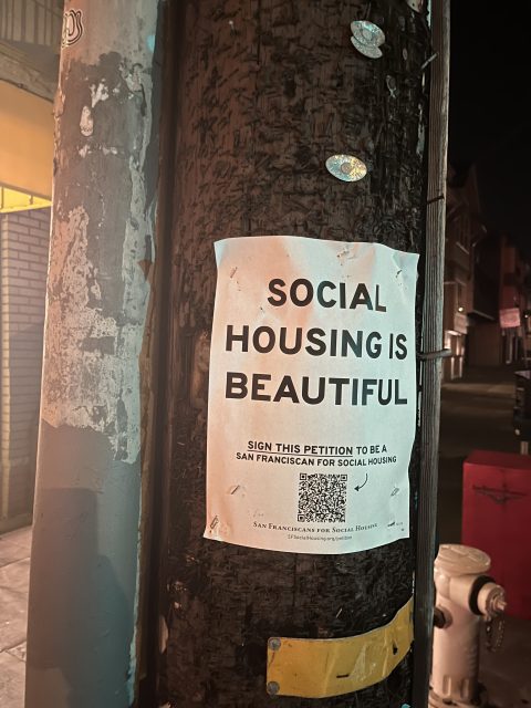 A paper sign reading "Social Housing Is Beautiful" is taped to a utility pole at night, with a QR code and petition link below the main message.
