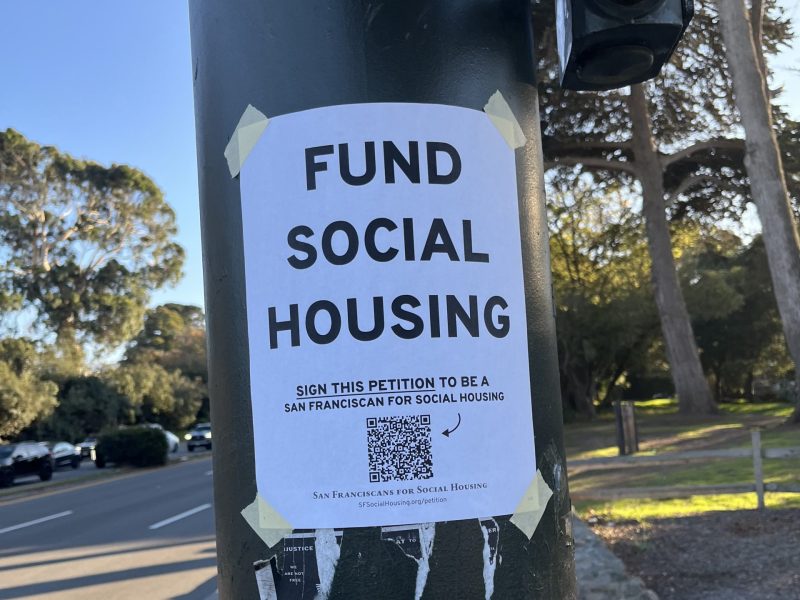 A flyer taped to a pole reads "Fund Social Housing" and urges people to sign a petition, with another notice below about public transit funding. Trees and a roadway are in the background.