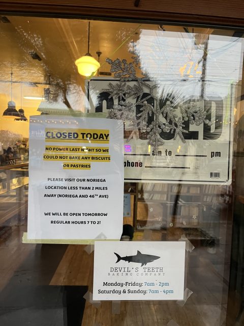 A bakery window displays a "Closed Today" sign due to a power outage, with business hours and an alternative location listed below. Indoor lights and reflections are visible.
