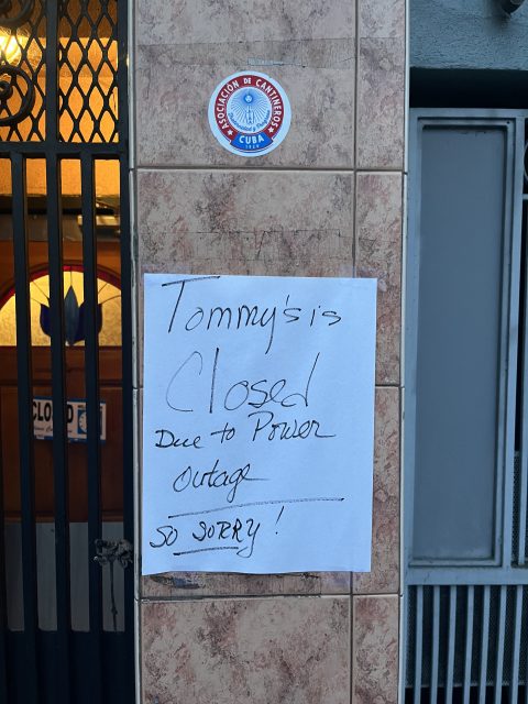 A handwritten sign on a tiled wall reads, "Tommy's is closed due to power outage. So sorry!" next to a door and a blue and red circular sticker above.