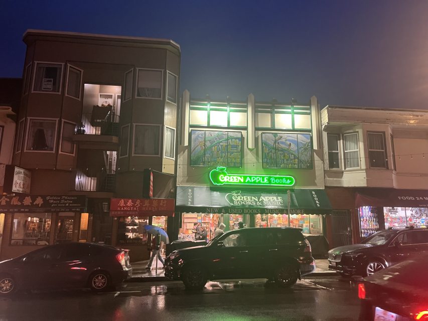Street view of Green Apple Books & Music at night, with neon sign lit and people walking past on wet sidewalk among parked cars.