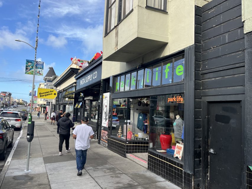 View of a city street with people walking on the sidewalk past shops, including a store called "Park Life" with large windows and a colorful sign.