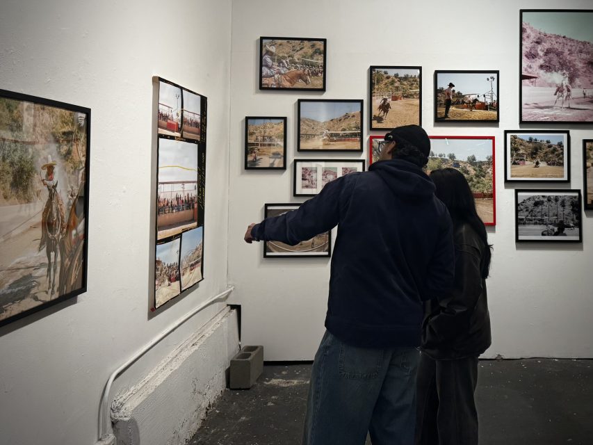 Two people stand in an art gallery looking at framed photographs of outdoor scenes by a Mexican American artist, displayed on white walls.