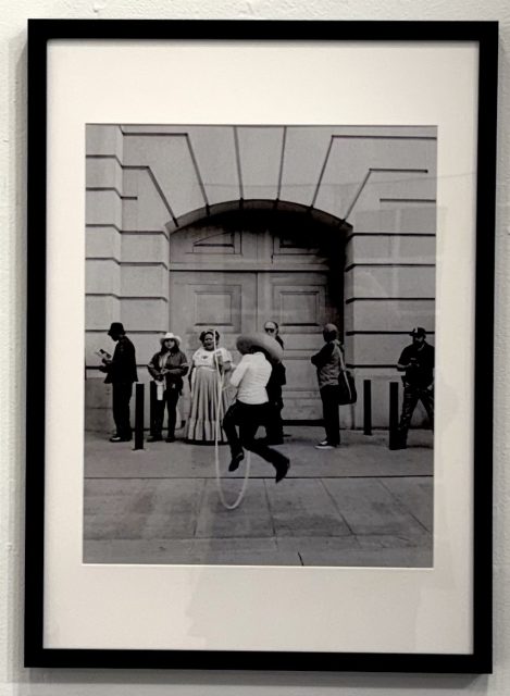 Two black and white framed photographs on a white wall; one features a Mexican American holding a U.S. flag, the other shows a group standing outside a building.