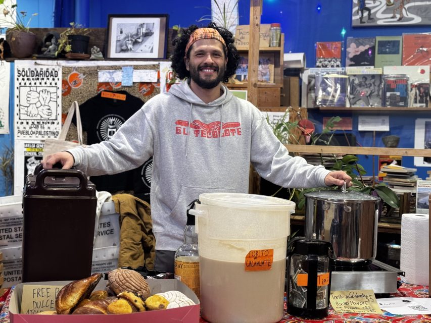 A man stands behind a table with sweet bread, a large container of drink, a pot, and cups, indoors at a community event or market.