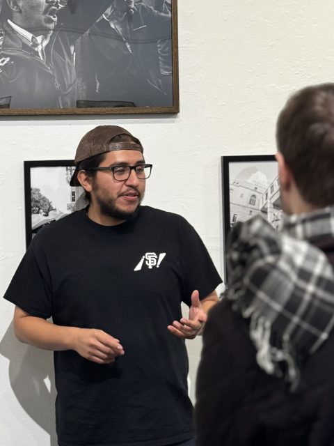 A man wearing glasses and a cap speaks to another person in front of framed photographs on a gallery wall.