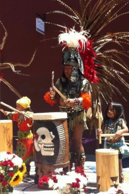 Three people in traditional Aztec attire stand behind decorated drums, performing outdoors among flowers. The central adult wears a large feathered headdress.