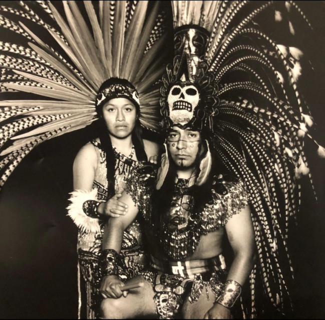 Two people wearing elaborate Aztec-inspired headdresses and traditional attire pose for a black and white portrait, with serious expressions and detailed accessories.