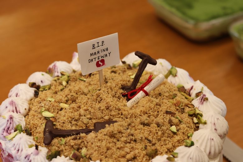 A cake decorated with crumbled topping, pistachios, chocolate shovels, a scroll, and a sign reading "R.I.P. Marina Gateway" on a wooden surface.