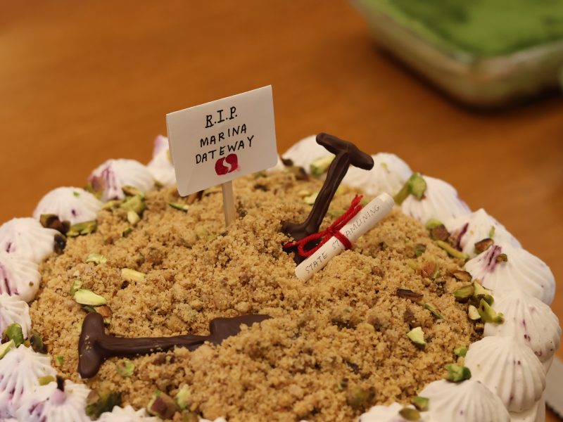 A cake decorated with crumbled topping, pistachios, chocolate shovels, a scroll, and a sign reading "R.I.P. Marina Gateway" on a wooden surface.