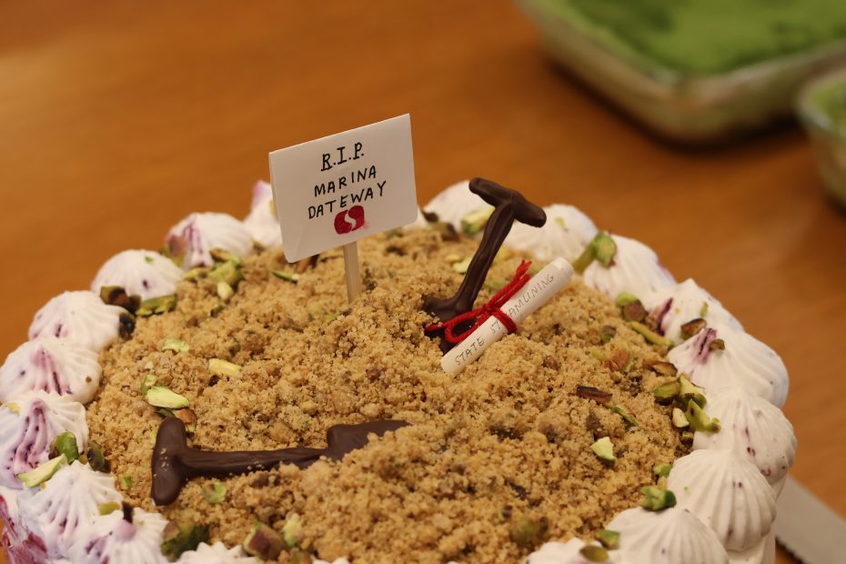 A cake decorated with crumbled topping, pistachios, chocolate shovels, a scroll, and a sign reading "R.I.P. Marina Gateway" on a wooden surface.