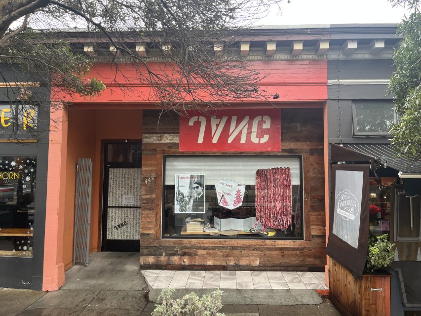 Street view of the front of a studio with a red and white sign, wood paneling, and display items in the window, including books and large prints.
