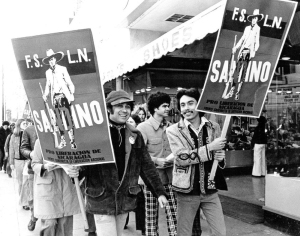 Group of people walking on a city sidewalk holding large posters with the text "F.S.L.N. SANDINO" and an image of a man in a hat and boots.
