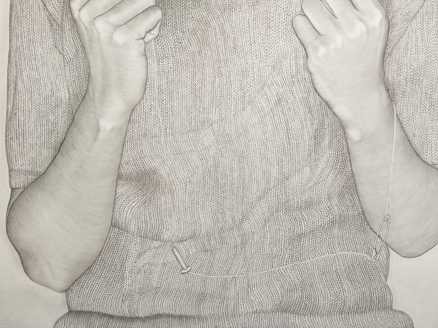 A person wearing a knitted sweater holds up both hands near their face, with a thin wire or string draped across their torso and attached to their right sleeve.