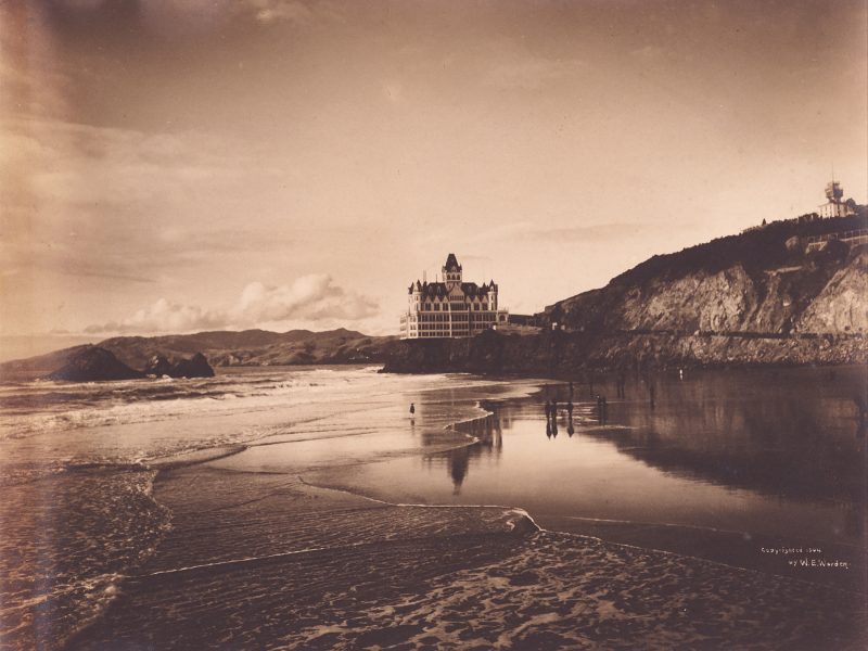 Sepia-toned photo of a large Victorian-style hotel on a cliff overlooking a beach with shallow waves and a few people near the water’s edge.