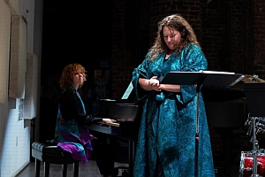 A woman plays piano while another woman stands beside a music stand, both wearing teal robes, on a dimly lit stage.