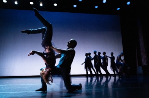 A group of dancers perform on stage under spotlights, with one dancer holding another upside down in an acrobatic pose in the foreground.