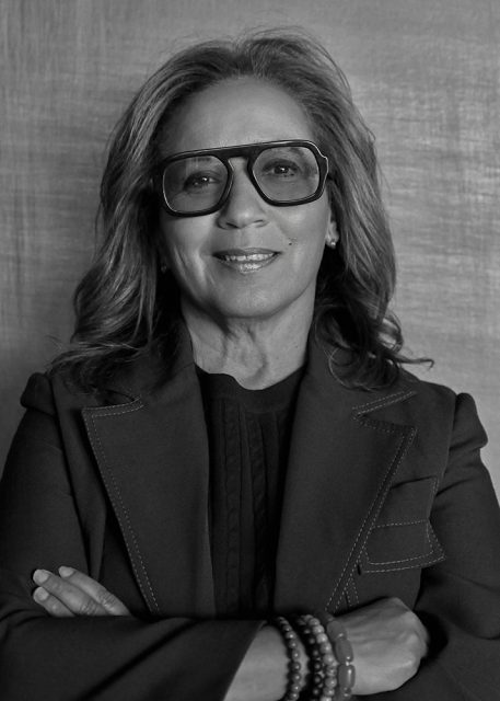 A woman with long hair and glasses, wearing a dark suit and beaded bracelets, stands with arms crossed in front of a plain textured background.