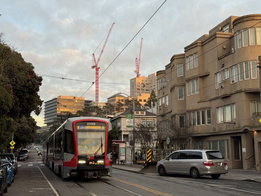 A red and white light rail train travels down a street lined with parked cars and beige buildings, with construction cranes visible in the background.