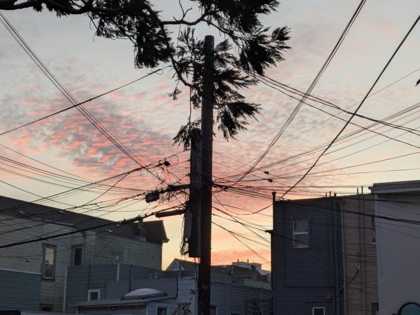 A utility pole with numerous tangled wires is set against a backdrop of buildings and a colorful sunset sky with scattered clouds.